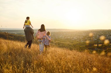Mutlu bir aile gün batımında tepenin yamacında yürüyor, altın çayır boyunca, arka manzara. Ebeveynler ve çocuklar birlikte yürüyüşten ve özgürlükten hoşlanırlar, baba kızını omuzlarında taşır, anne oğlanı elleriyle tutar.