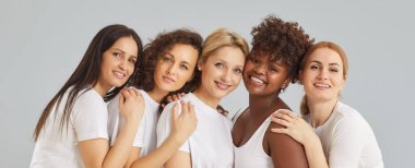 Stüdyonun gri arka planında izole edilmiş beyaz elbiseli ve kameraya bakan neşeli, çeşitli kadın arkadaşlarının yakın plan portresi. Çeşitlilik, destek ve takım konsepti. Pankart.