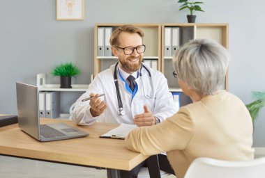 Yaşlı kadın hastane ziyareti sırasında erkek doktorla sağlık sorunlarını tartışıyor, doktor yaşlı hastayı dikkatle dinliyor, güvence veriyor ve tedavi planını anlatıyor