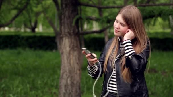 Ganska ung flicka lyssnar musik i hörlurar i parken — Stockvideo