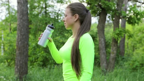 Женщина пьет воду после занятий спортом в лесу, затем смотрит в камеру и улыбается — стоковое видео