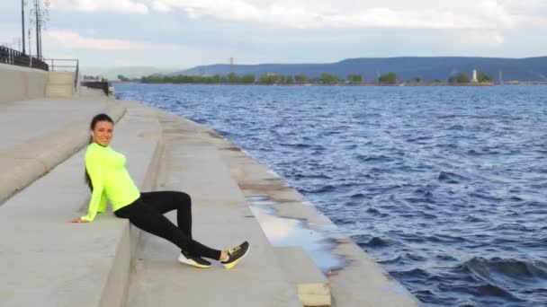 Chica morena caucásica en tela deportiva relajarse en terraplén escuchando música — Vídeo de stock