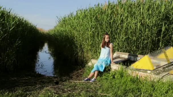 Молода дівчина-підліток сидить на старому човні біля озера сільський зелений очерет фону — стокове відео