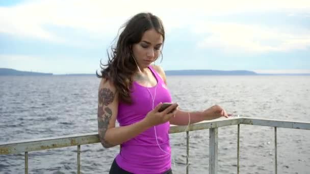 Fitness girl sur le remblai de la rivière. Détendez-vous en écoutant de la musique. 4K — Video