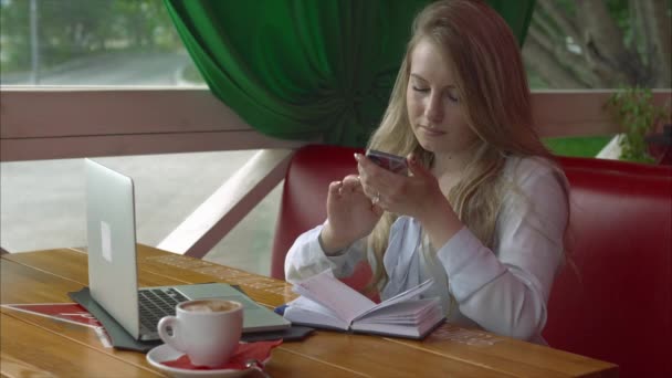 Γυναίκα των επιχειρήσεων σε μια καφετέρια που λειτουργεί με smartphone laptop και μικρόs φορήτοs — Αρχείο Βίντεο