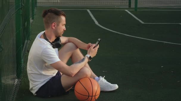 Σπορ άνθρωπος χαλαρώνει στο γήπεδο μπάσκετ δρόμο, χρησιμοποιώντας smartphone — Αρχείο Βίντεο