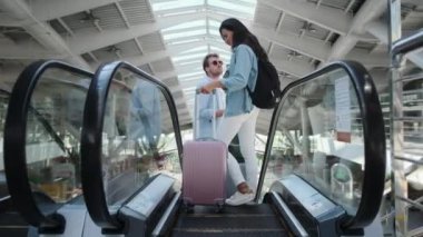 Tren istasyonunda yürüyen merdivende duran bavullu iki turist gülümsüyor.