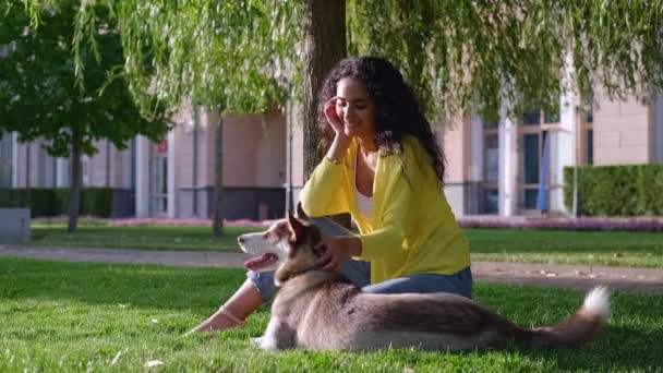 Отдых в парке с собакой, молодая женщина и ее питомец сидят на зеленой траве — стоковое видео