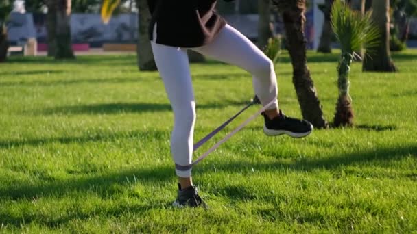 Entrenamiento al aire libre. Chica haciendo levantamientos de piernas con banda de resistencia — Vídeos de Stock