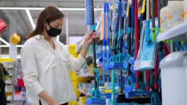 Yüz maskeli genç bir kadın mağazadan ev eşyalarıyla alışveriş yapıyor, paspas seçiyor.