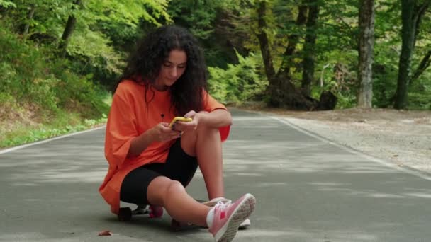 Deportiva chica usando móvil sentado en monopatín en el parque — Vídeos de Stock