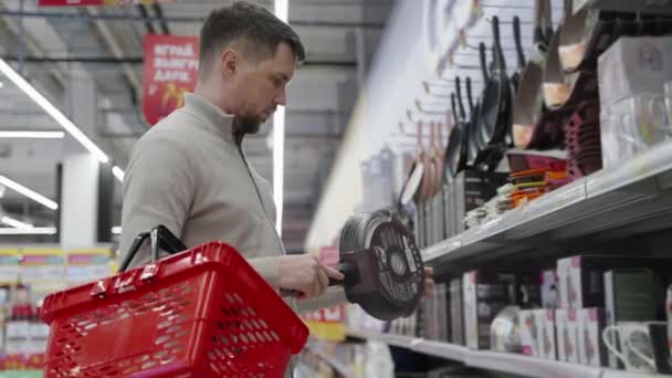 Hombre soltero está de compras en el hipermercado, la elección de sartén en el departamento de vajilla — Vídeo de stock