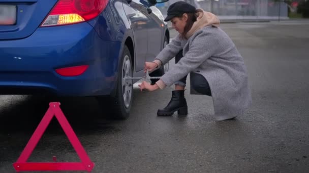 Young woman is trying to change broken wheel of her car on road, using balloon key — Stock Video