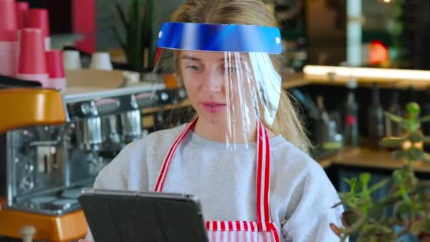 A kávézó női dolgozója tablettát használ, arcvédőt visel a koronavírus fertőzés ellen — Stock videók