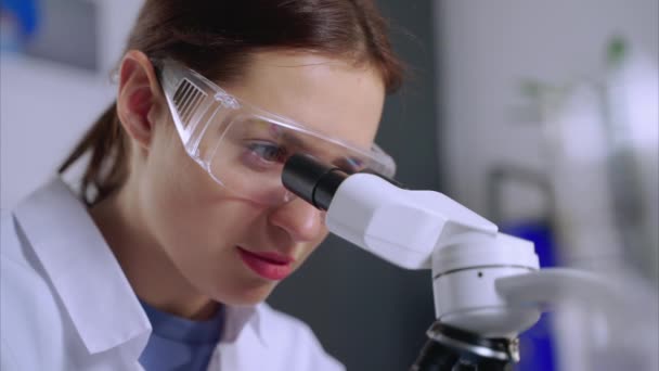 Jong vrouwelijk chemisch laboratorium kijkt in microscoop, close-up van het gezicht — Stockvideo