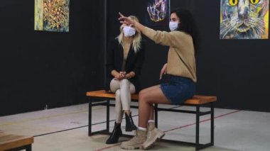 Maskeli iki kadın sanat uzmanı sanat müzesinde oturmuş resimlere bakıyorlar.