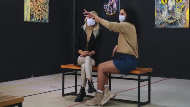 Dois apreciadores de arte do sexo feminino com máscaras estão sentados no hall do museu de arte e olhando para fotos — Vídeo de Stock