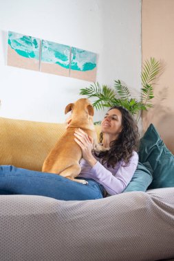 Young woman lying on a comfortable sofa, cuddling her small brown dog in a cozy home setting