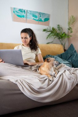 Freelancer working remotely from home office with her dog, using laptop computer on the sofa, enjoying comfortable lifestyle