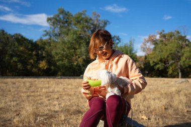 Güneşli bir doğa yürüyüşü sırasında küçük köpeğini sulayan bir kadın.
