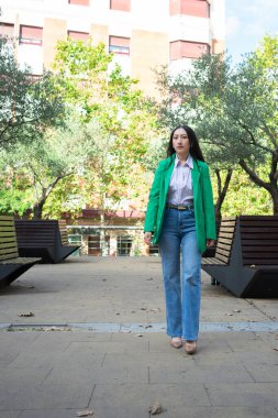 Genç bir kadın yeşil spor ceketiyle şehir parkında rahatça yürüyor.