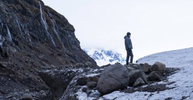 Buzul, kayalık bir dağ ve kışın karla kaplı zirveleri olan kayaların üzerinde duran adam.