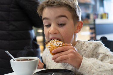 Çocuk tatlı bir hamur işi yiyor. Yanında da sıcak çikolata serpiştiriyor.