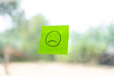 Şeffaf bir pencere camına yapışmış üzgün yüzlü yeşil yapışkan bir not.