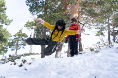 İki çocuk ormanda karlı bir günün tadını çıkarırken, biri neşeyle zıplıyor, diğeri izliyor.