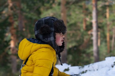 Sıcak kışlık giysiler giymiş genç bir çocuk karlı bir ormanda oynuyor, kış harikalar diyarının tadını çıkarıyor.