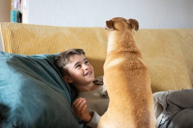Çocuk gülümsüyor, kanepede dinleniyor, köpeğinin onunla etkileşimine bakıyor.