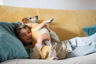 Mutlu çocuk evcil köpeğini kucaklıyor. Sarı koltukta mavi yastıkla dinleniyor.