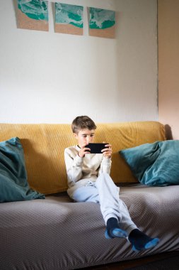 Çocuk oturma odasındaki kanepede online oyun oynamak için akıllı telefonu kullanırken konsantre oluyor.