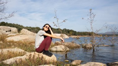 Genç bir kadın gölün kenarındaki bir kayanın üzerinde güneşli bir günün tadını çıkarıyor.