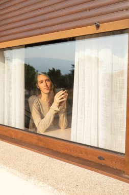 Kadın evde dinleniyor, bir fincan kahvenin tadını çıkarıyor ve pencereden dışarı bakıyor.
