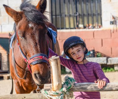 Kasklı küçük bir çocuk, kahverengi bir atın yanındaki tahta çitlere yaslanıyor.