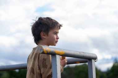 Genç bir çocuk dışarıda bulutlu bir gökyüzüne bakıyor, elinde bir aperatif, profil görüntüsü var.