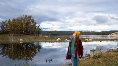 Yansıtıcı bir gölün kenarında duran kadın bulutlu sonbahar gökyüzüne bakıyor.