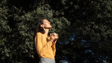 Kadın dışarıda duruyor, gözlerini kapatıyor ve sıcak bir bardak tutarken güneşi hissediyor.