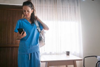Önlüklü kadın hemşire işe hazırlanıyor, akıllı telefon tutuyor ve sırt çantası taşıyor.