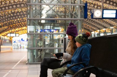 Anne ve oğul tren istasyonunda bir bankta oturup akıllı telefon kullanarak trenlerini bekliyorlar.