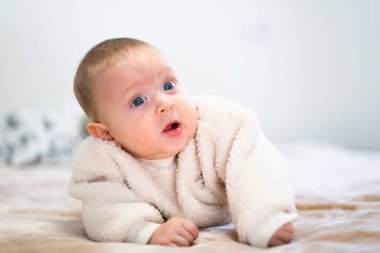 Mavi gözlü sevimli bebek, yumuşak bir battaniyenin üzerine uzanmış başını kaldırıyor ve merakla bakıyor.