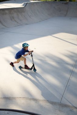 Güneşli bir günde kaykay parkında kasklı bir çocuk scooter kullanıyor.