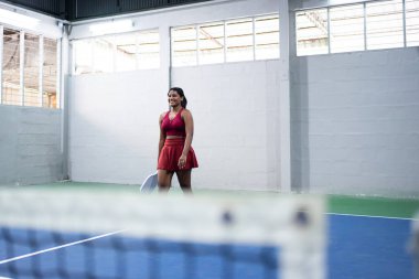 Gülümseyen raket tenisçisi elinde raketle iç sahada yürüyor.