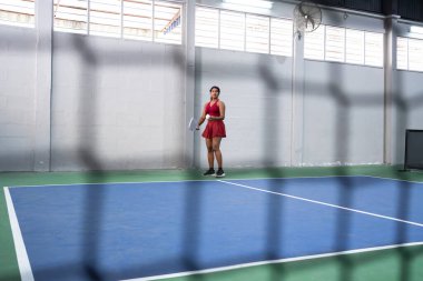 Pickleball oynayan genç bir kadın, raket tutuyor ve kapalı sahada top oynamaya hazırlanıyor.