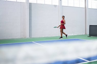 Genç bir kadın, kapalı alanda top oynuyor, raket tutuyor ve sahada yürüyor.