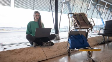 Havaalanı penceresinde dizüstü bilgisayar kullanan, uzaktan kumandalı, uçuş beklerken çalışan bir kadın.