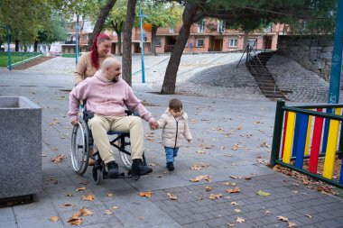 Aile birlikte yürüyor, baba tekerlekli sandalyede çocuğun elini tutuyor.