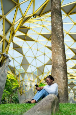 Öğrenci akıllı telefon uygulaması kullanıyor. Parktaki parlak sarı metal ve cam kubbenin altında oturuyor.