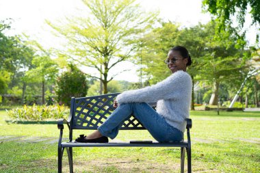 Parktaki bankta oturan genç siyahi kadın güneşli bir günün tadını çıkarıyor.